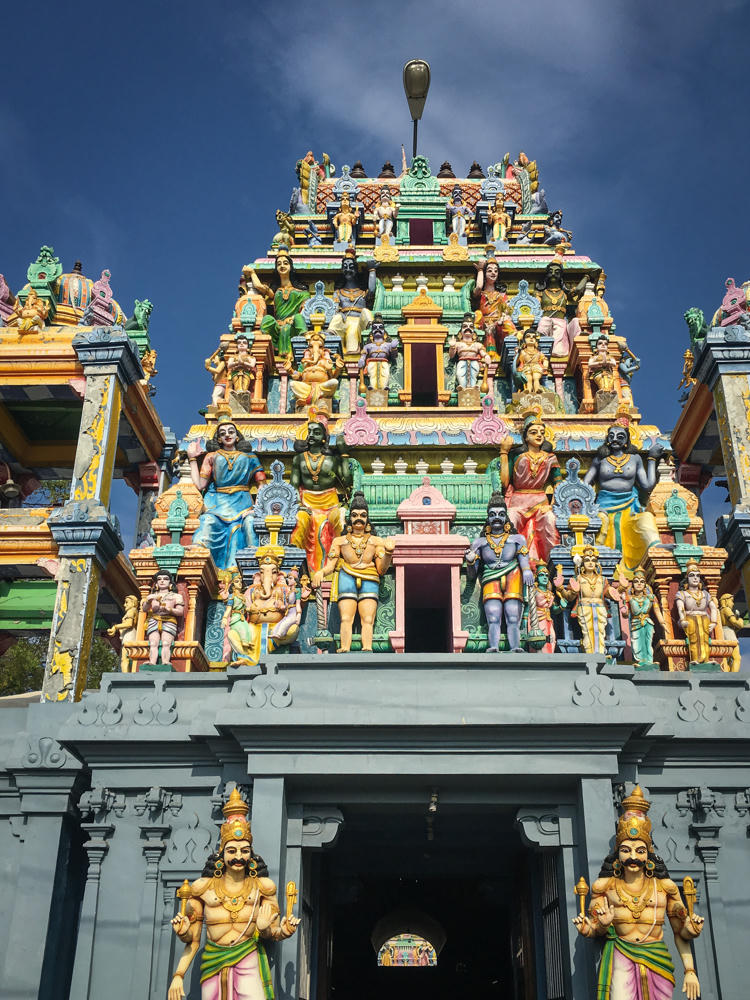 A towering, brightly colored Hindu temple gopuram covered in intricate sculptures of deities and figures, viewed from below against a deep blue sky. Two large golden guardian statues flank the dark entrance gateway at the base.