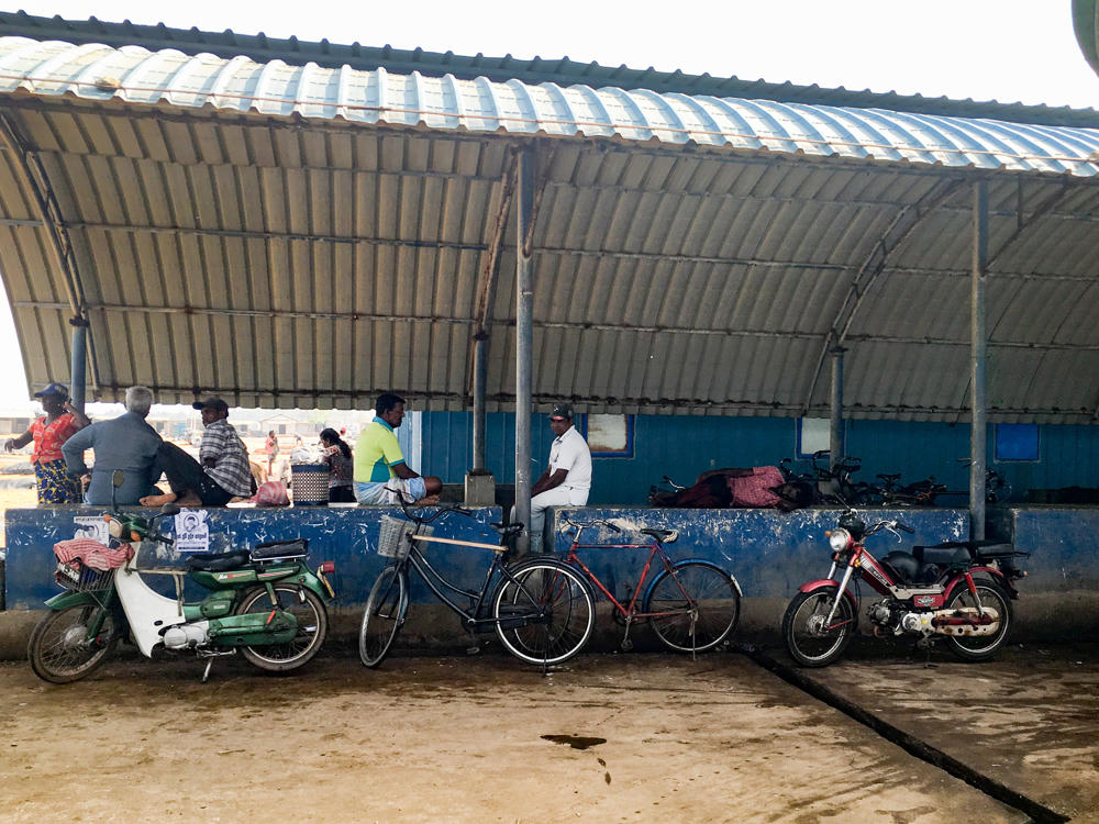 A group of people sit and stand along a long blue painted concrete ledge under a curved corrugated metal roof at an open-sided market hall, with two mopeds and two bicycles parked against the ledge on the concrete floor in front of them.