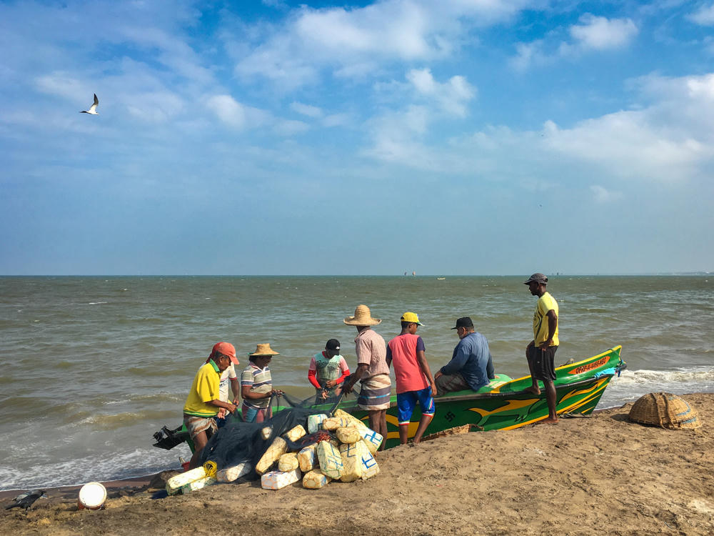 A group of fishermen in colorful shirts and sun hats gather around a green and yellow fishing boat on a sandy beach beside the sea, with floats, nets, and woven baskets piled on the sand and a bird flying in the cloudy blue sky.