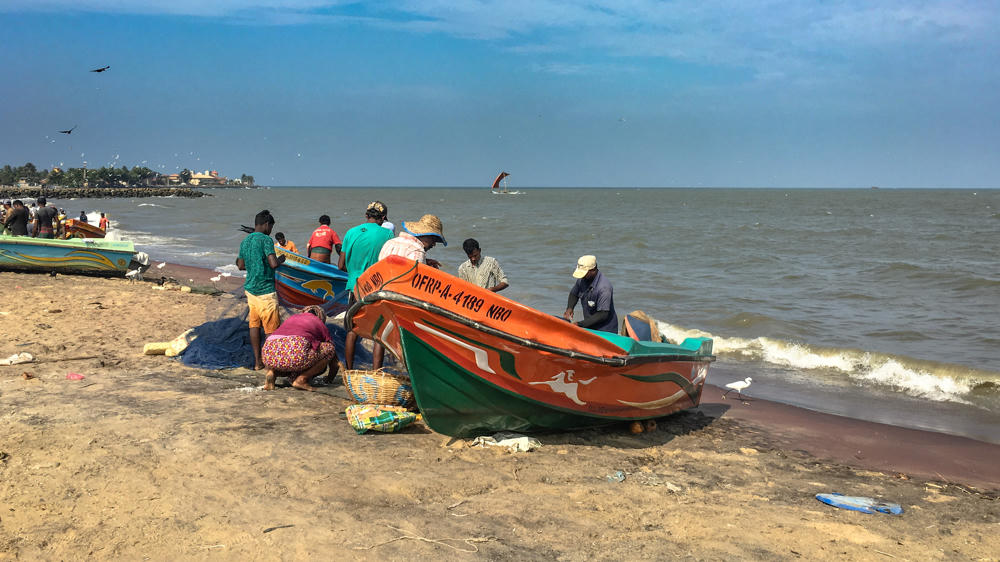 A group of fishermen gather around colorful wooden fishing boats on a sandy beach, with one bright orange and green boat marked 'OFRP-A-4189 NBO' in the foreground. Beyond the calm sea a sailing boat is visible, with birds in the hazy blue sky and a white egret standing at the water’s edge.