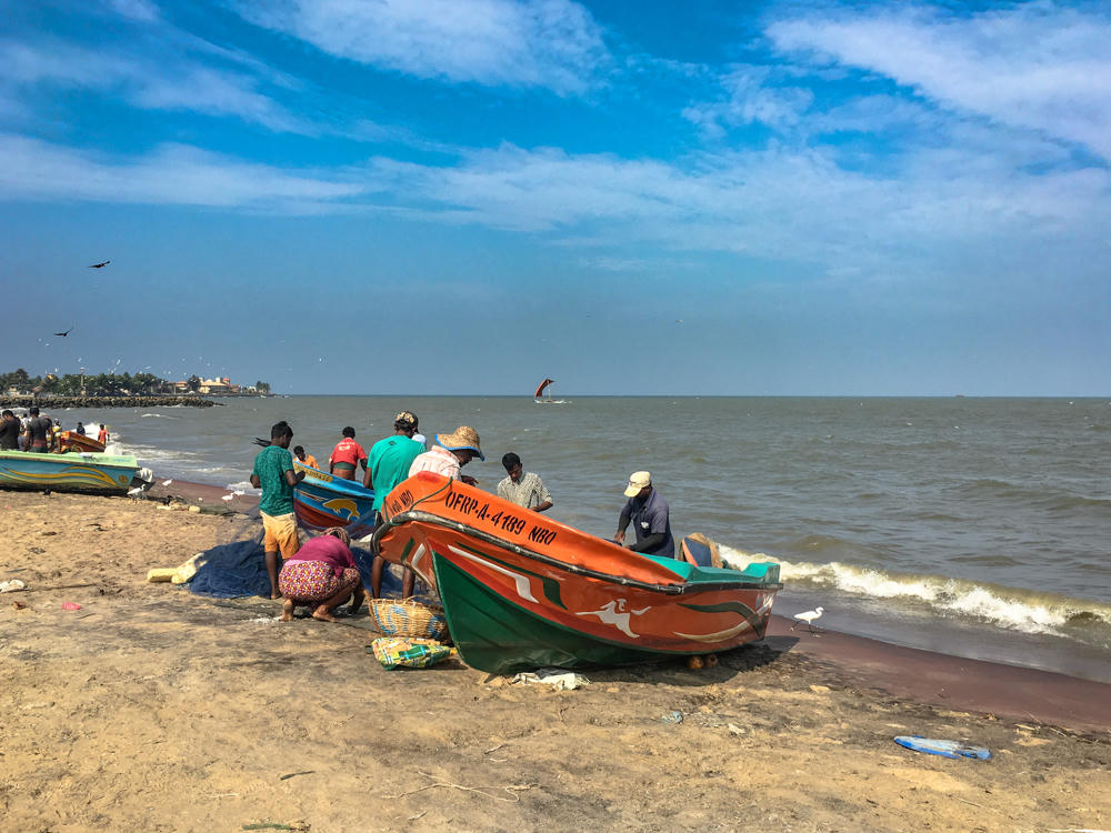 A group of fishermen gather around colorful wooden fishing boats on a sandy beach, with one bright orange and green boat marked 'OFRP-A-4189 NBO' in the foreground. Beyond the calm sea a sailing boat is visible, with birds in the hazy blue sky and a white egret standing at the water’s edge.