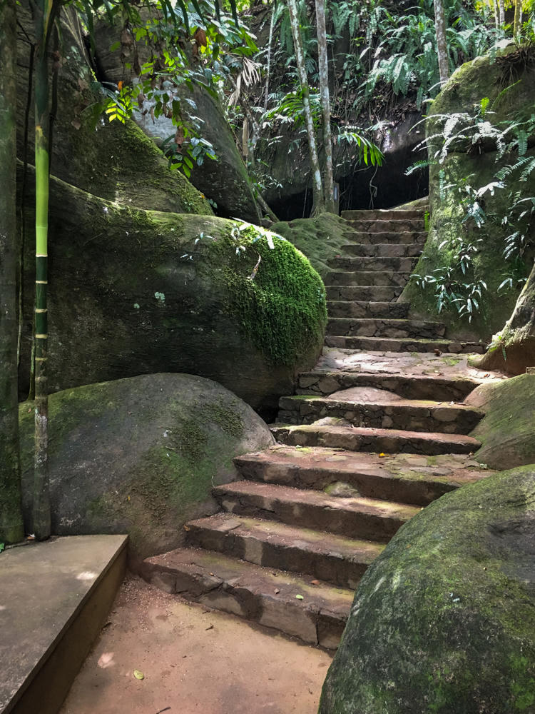 A winding stone staircase climbs between large moss-covered boulders in a lush tropical forest, with bamboo stalks and dense green ferns and foliage surrounding the path at Boulder Garden in Kalawana, Sri Lanka.