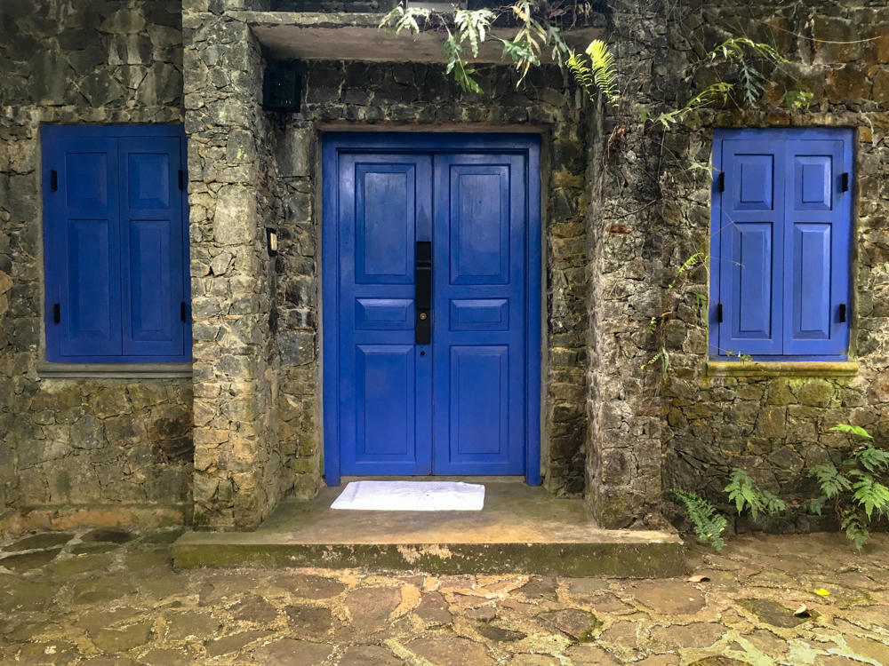 A rough stone wall facade with a bright blue wooden double door flanked by two matching blue shuttered windows, with a white doormat on the concrete step and ferns and foliage growing along the wall, set on a flagstone path.