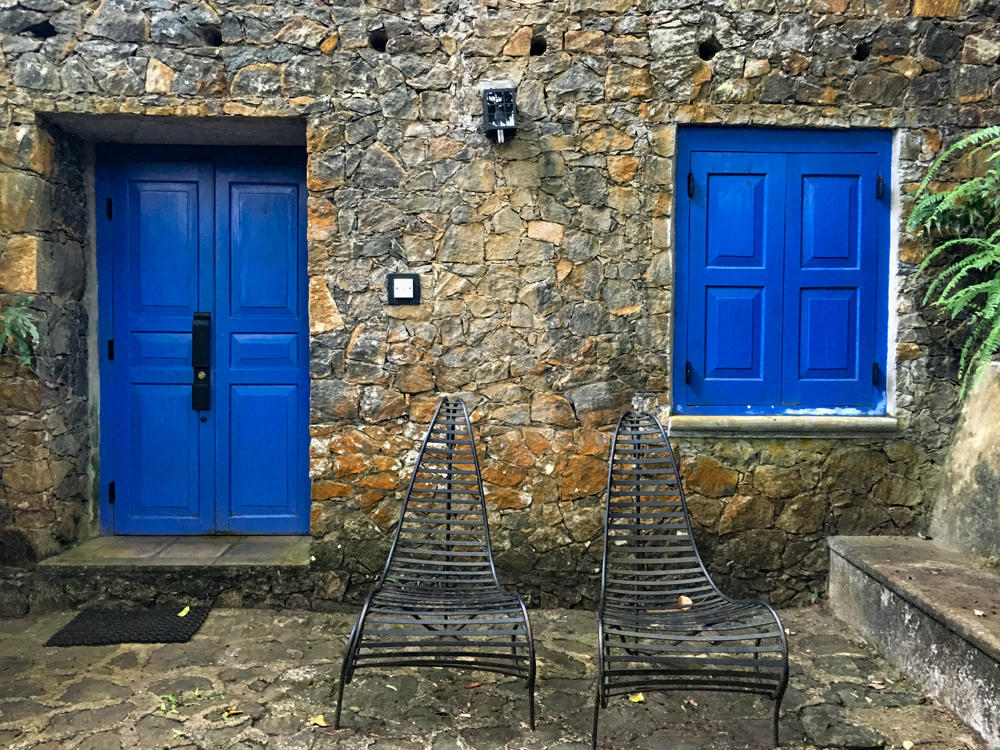 Two black wrought-iron lounge chairs sit on a cobblestone surface against a rustic stone wall, which has a bright blue paneled door on the left and a matching blue shuttered window on the right. Green ferns grow at the edges and a small black lantern is mounted on the wall.