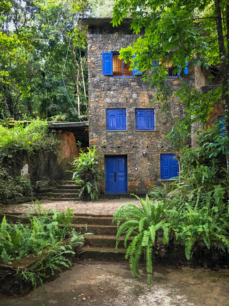 A three-story stone building with bright blue painted doors and shuttered windows, nestled in dense green rainforest with lush ferns and trees surrounding it. Stone steps lead up toward the building's blue front door in the overgrown jungle setting.