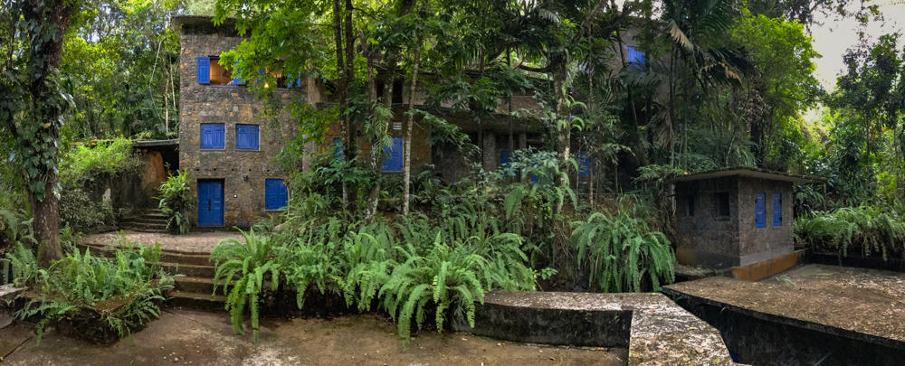 A multi-story stone building with bright blue shuttered windows and doors set deep within lush tropical rainforest, surrounded by tall trees, ferns, and dense green foliage. A smaller stone outbuilding stands to the right, with stone steps and a paved area in the foreground.