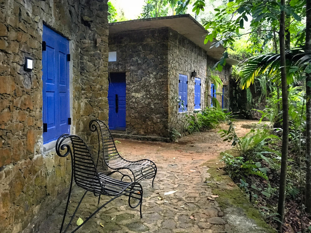 A rustic stone building with bright cobalt-blue painted doors and shutters sits along a cobblestone and dirt pathway, surrounded by lush green tropical foliage including palms and ferns. A black wrought-iron chair with a curled, decorative frame stands empty in the foreground.