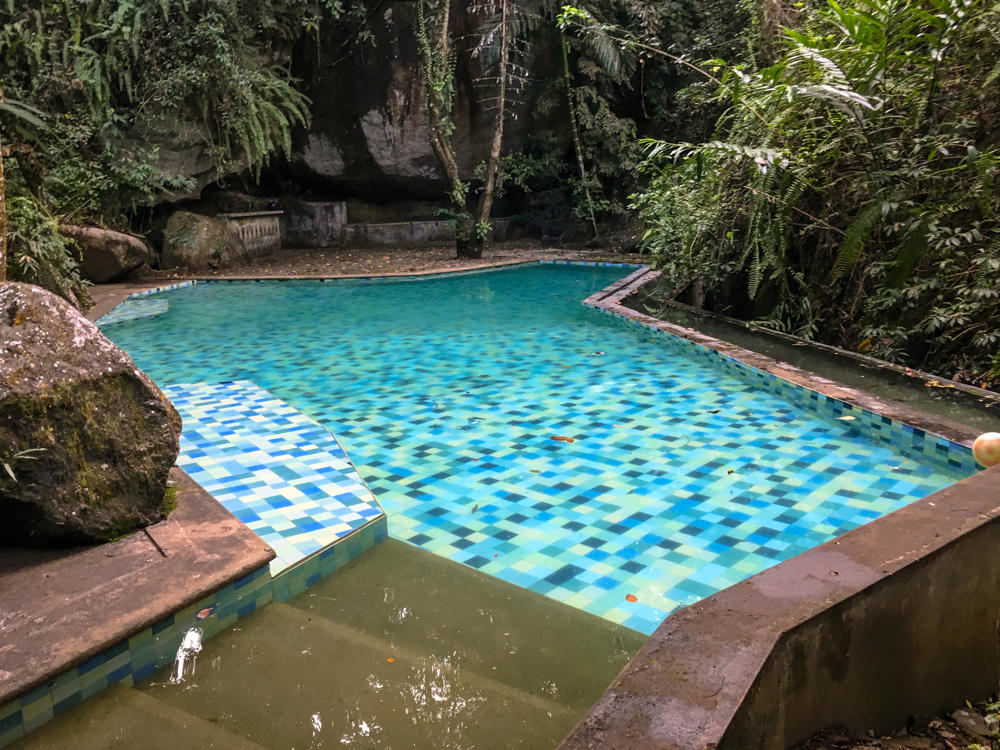 A blue-and-green mosaic-tiled swimming pool set into a lush rainforest, with clear turquoise water giving way to a murky greenish shallow end in the foreground. Large moss-covered boulders, a dark rock overhang, and dense tropical greenery surround the pool.