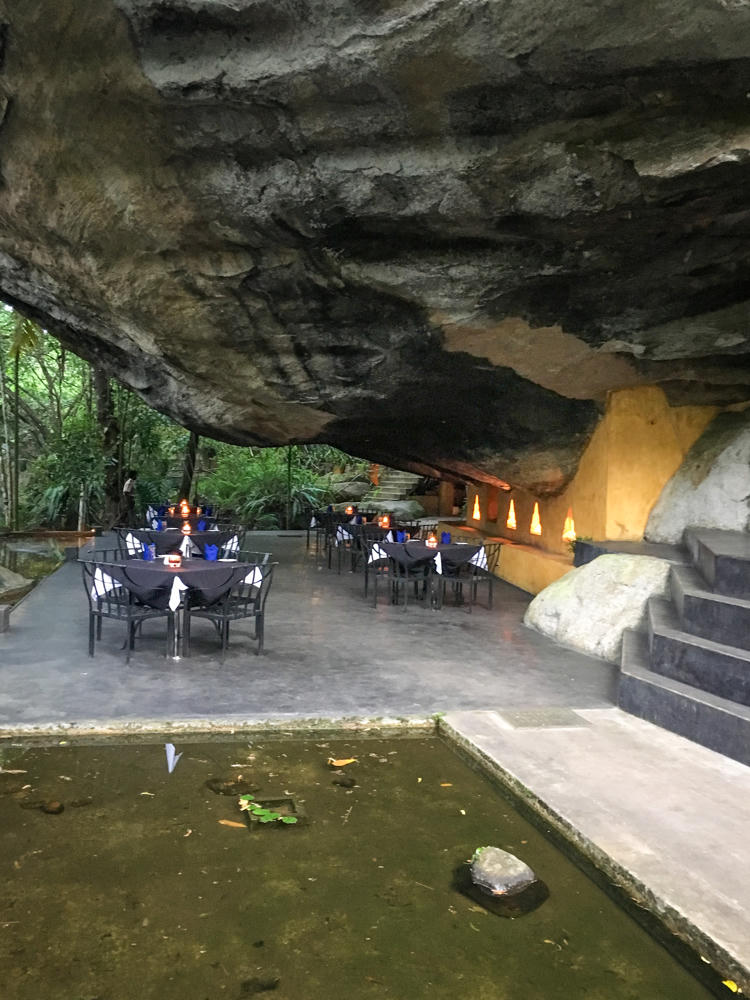 An open-air restaurant set beneath a massive overhanging boulder, with dark-clothed tables set with blue napkins and small candle lamps arranged on a polished grey stone floor. Lush green jungle is visible to the left, candle lamps line the rock wall, stone steps rise on the right, and a shallow reflecting pool sits in the foreground.