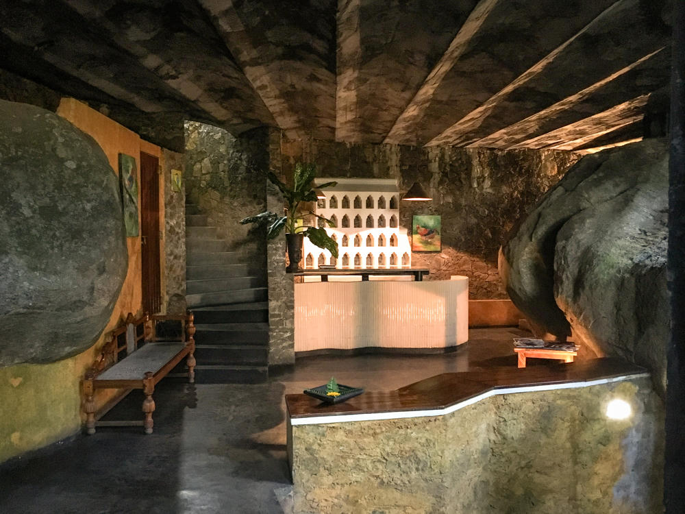 A dimly lit interior reception area built into and around enormous natural boulders that form the walls and partly the ceiling, with a curved tiled reception desk, a tall leafy potted plant, a wooden bench against a boulder, and a stone staircase leading up at left. A small white architectural model and framed paintings decorate the space, and polished stone surfaces reflect the warm lighting.