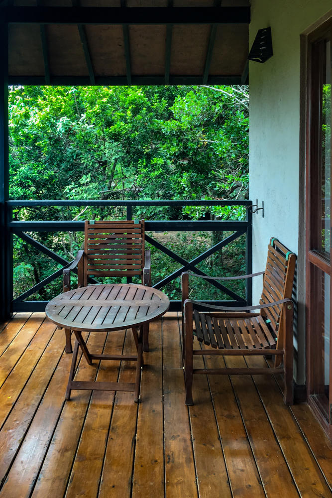 A wooden balcony with two slatted teak armchairs and a round wooden table, set under a sloped roof with dark beams and overlooking dense green forest foliage. A black metal railing borders the deck, and french doors with glass panes open into the room on the right.