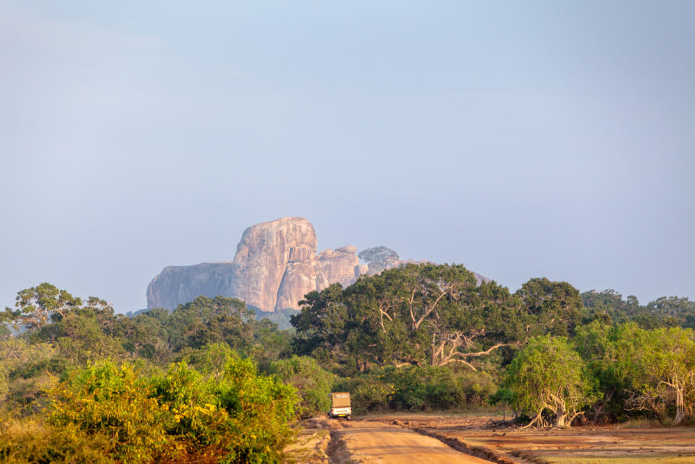 A large rounded rock outcrop rises above dense green woodland under a hazy pale blue sky, with a dirt track running through scrubland in the foreground where a small truck is parked.