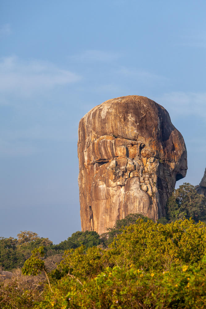 A massive weathered granite boulder rising above dense green scrubland under a pale blue sky, its rounded, eroded surface forming craggy ridges that suggest a face in profile. A smaller rock formation is visible to the right, partly hidden by trees.