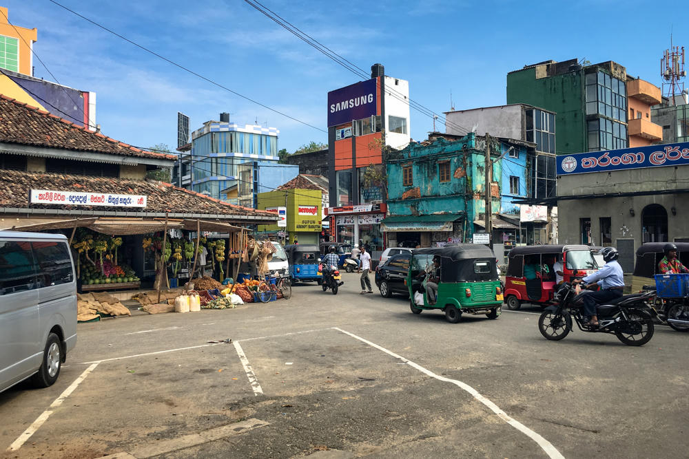 A busy street scene in Galle, Sri Lanka, with green, red, and white auto rickshaws (tuk-tuks), a motorcyclist, and a parked silver van moving across a wide paved junction. The roadside is lined with weathered buildings, shop signs in Sinhala, a Samsung billboard, and an open-air fruit and vegetable stall with hanging bananas under a clear blue sky.