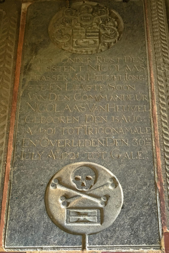 A weathered stone gravestone with a carved coat of arms at the top and a skull-and-crossbones emblem below, surrounded by a rope-like decorative border. The Dutch inscription commemorates Nicolaas Brasser van Heuvel, born in Trincomalee in 1701 and died in Galle in 1721.