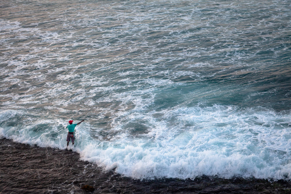 A lone fisherman in a teal shirt and red cap stands on dark rocks at the water’s edge, casting into rough, foaming surf that fills most of the frame with churning blue-green sea.