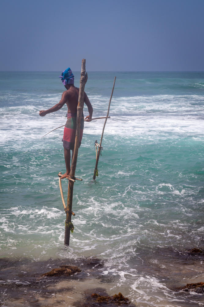 A stilt fisherman in Koggala, Sri Lanka, perched on a thin wooden pole driven into the shallow sea, balancing with his feet on a crossbar and holding a fishing rod. He is bare-chested, wearing a red and green sarong and a blue head wrap, facing the turquoise water with breaking white waves and a hazy horizon behind him.