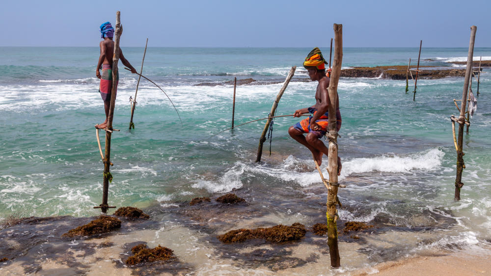 Two stilt fishermen perched on tall wooden poles set into shallow turquoise water along a rocky coastline, each holding a slender fishing rod, with breaking waves and a hazy sea horizon behind them. One man stands wearing a blue head wrap and a pink wrap, while the other sits on a crossbar wearing a colorful headscarf and orange shorts.