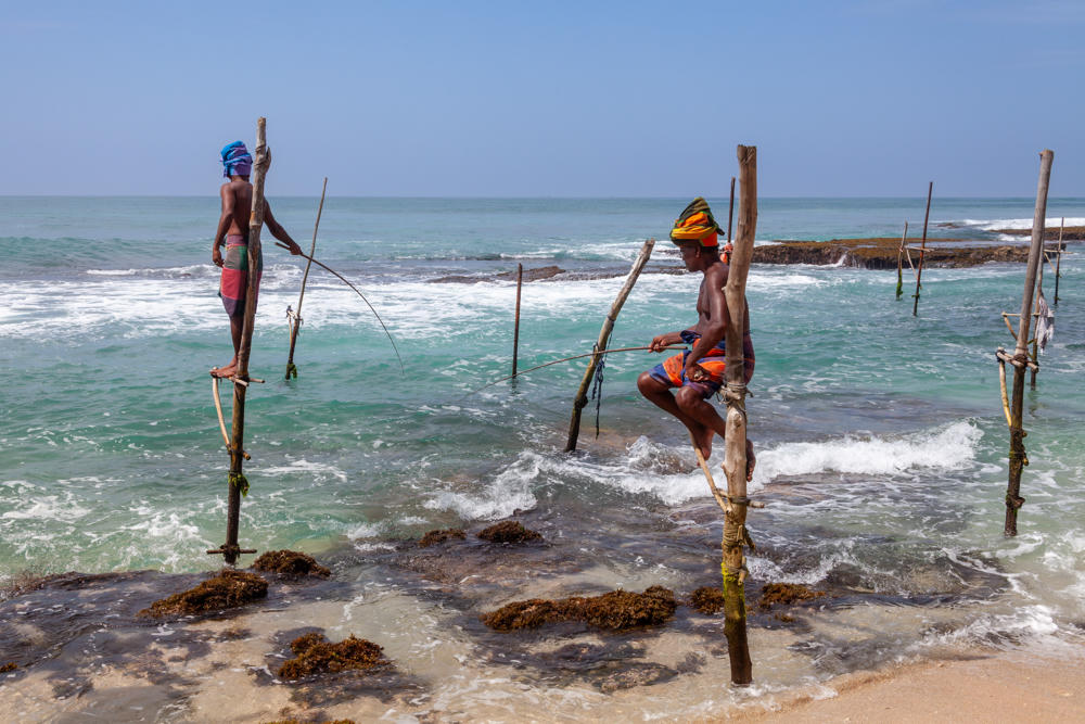 Two stilt fishermen perched on tall wooden poles set into shallow turquoise water along a rocky coastline, each holding a slender fishing rod, with breaking waves and a hazy sea horizon behind them. One man stands wearing a blue head wrap and a pink wrap, while the other sits on a crossbar wearing a colorful headscarf and orange shorts.
