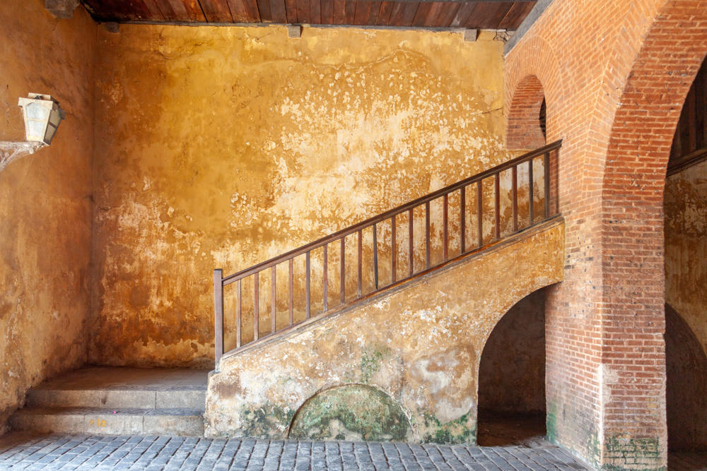 A weathered interior space at Galle Fort with ochre-colored, mottled plaster walls, a wooden-railed staircase rising to the right beneath a brick archway, and a stone-paved floor. A metal lantern is mounted on the left wall, and the ceiling is made of dark wooden beams and planks.