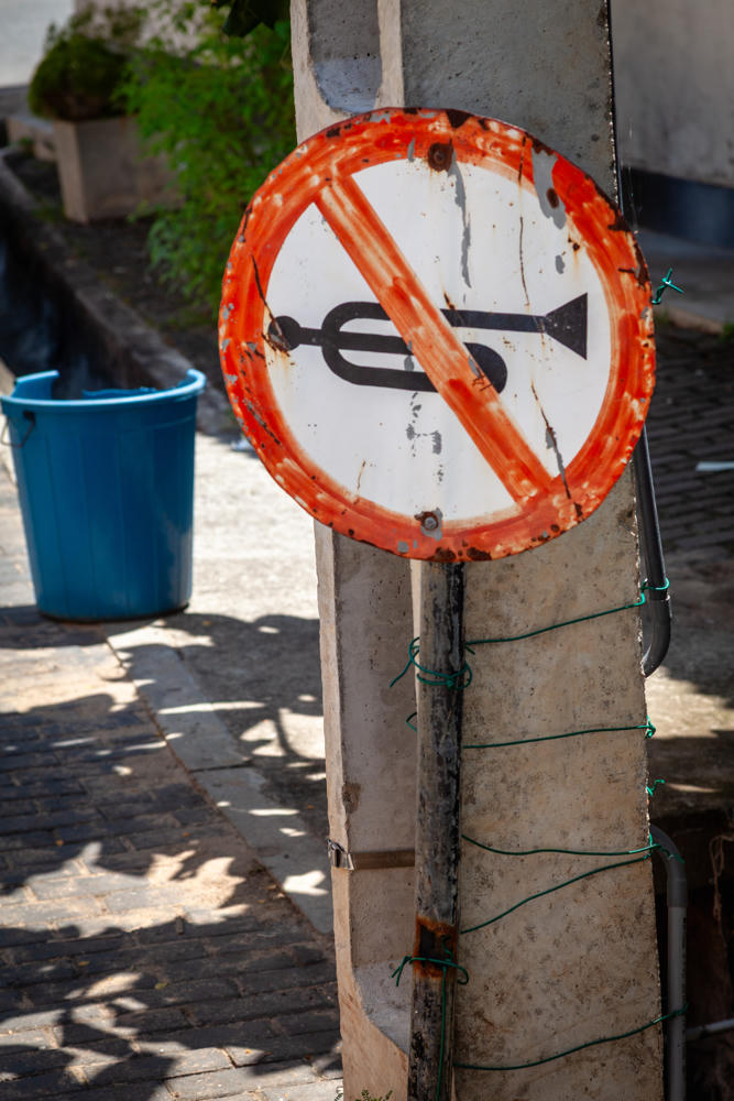 A weathered round “no honking” traffic sign with a black trumpet symbol crossed out, its faded orange-red border rusted and peeling, mounted on a concrete post wrapped with green wire. A blue plastic bin sits on a sunlit stone pavement in the background, with a planter of greenery nearby.
