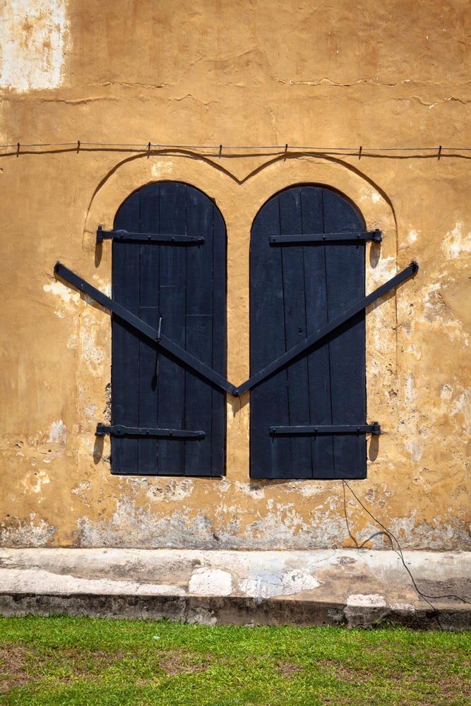A pair of arched black wooden shutters with iron hinges and diagonal braces set into a weathered ochre-yellow plaster wall, the two arches together forming a heart-like shape, with a strip of grass below at Galle Fort in Sri Lanka.