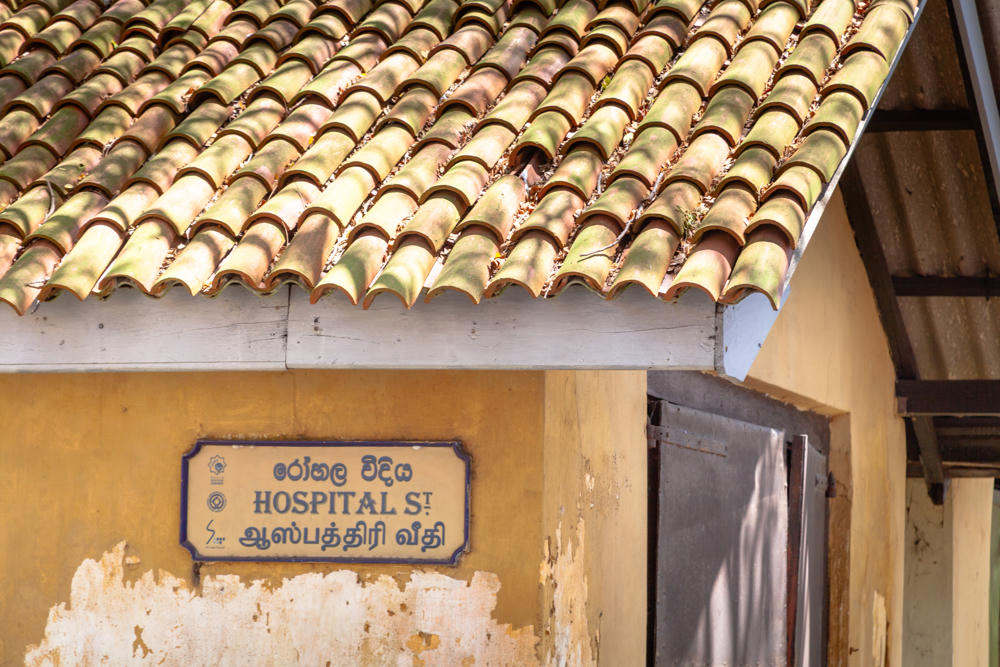 A trilingual street sign reading “Hospital Street” in English, Sinhala, and Tamil mounted on a weathered yellow-ochre wall with peeling plaster, beneath a terracotta tiled roof. A dark doorway opens to the right in the colonial-era building in Galle Fort, Sri Lanka.