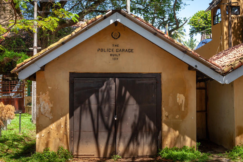 An old single-story garage with a yellow-ochre stucco facade and red tiled gable roof, its front gable wall painted with an emblem and the words “The Police Garage Built 1917” above a padlocked pair of dark metal doors. Lush green trees and grass surround the weathered building under bright daylight.