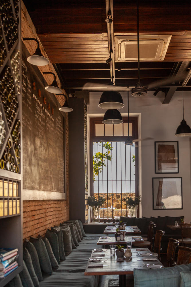 The dimly lit interior of a restaurant with a long row of tables flanked by grey cushioned bench seating along an exposed brick and chalkboard wall, with wine bottles stored in a wall rack, black pendant lamps hanging from a high wooden-beamed ceiling, and tall barred windows letting in daylight.