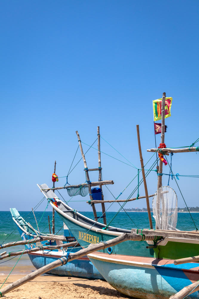 Brightly painted wooden outrigger fishing boats with tall bamboo masts and rigging rest on a sandy beach beside the turquoise sea, one bearing the name “MAHEESHA” in yellow lettering. A small Sri Lankan flag and colorful flower garlands hang from the rigging, with cast nets and a blue bucket aboard, under a clear blue sky.