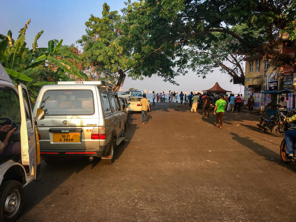 A waterfront road in Vypin, Kerala lined with parked vehicles including a silver Toyota SUV with a yellow Indian number plate and white vans, with people and motorbikes gathered near the shore in the late afternoon. Lush banana plants and leafy trees frame the scene, and a yellow building marked “Hotel” stands on the right with the sea visible in the distance.