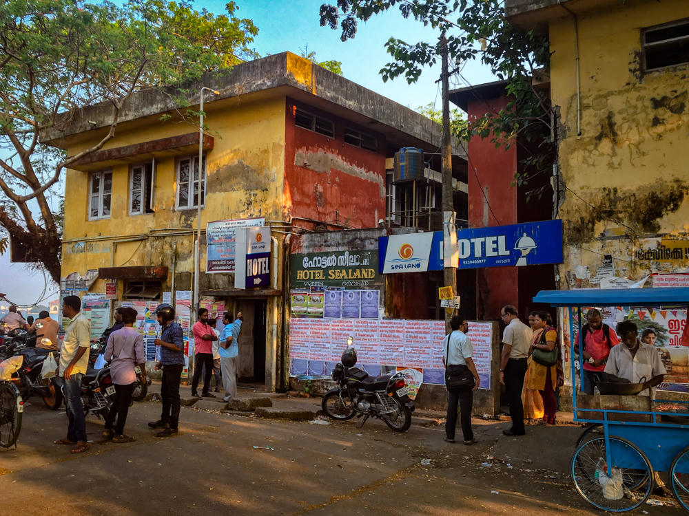 A weathered Indian street scene outside Hotel Sealand, with peeling yellow and red building facades covered in posters and blue ‘HOTEL’ signs. People stand and walk along the road past parked motorcycles, with a blue street-food cart and a vendor cooking on the right, beneath leafy trees in warm late-afternoon light.