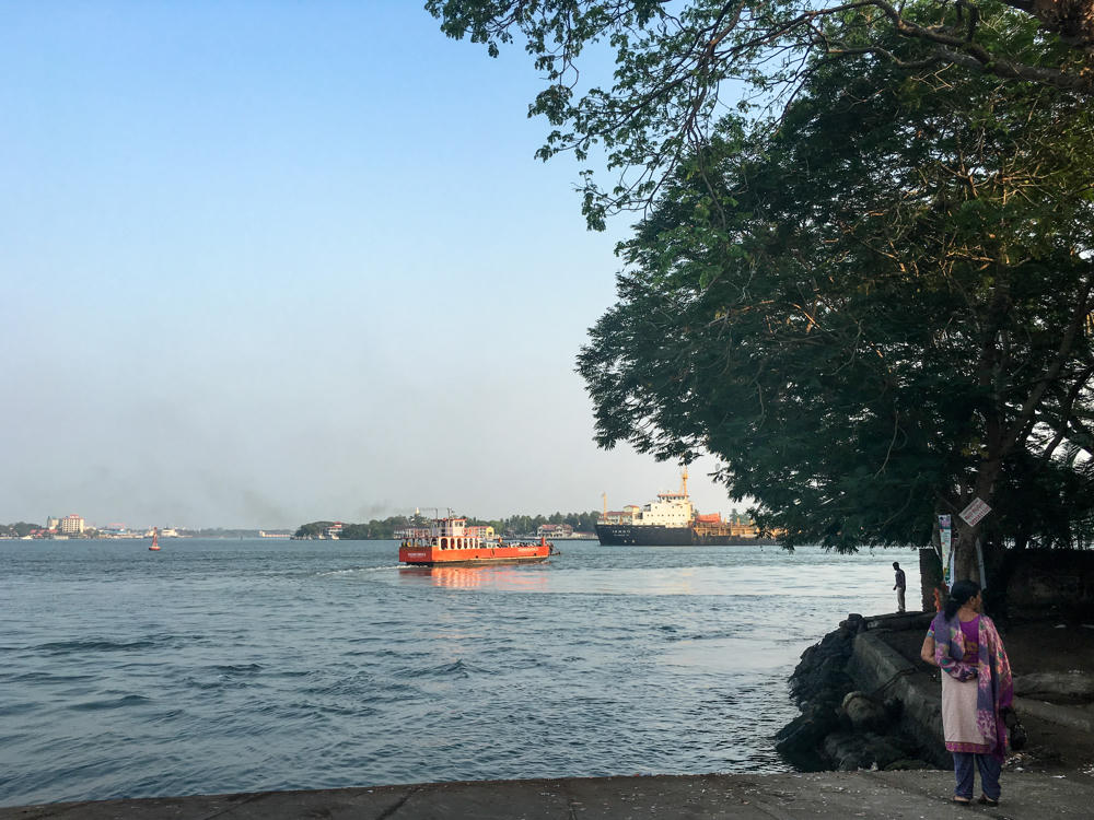 A waterfront scene in Vypin, Kerala, where a woman in a purple and pink salwar kameez stands on a rocky embankment beneath a large leafy tree on the right. An orange ferry crosses the calm water in the distance, with a large cargo ship and a hazy shoreline visible under a pale sky.