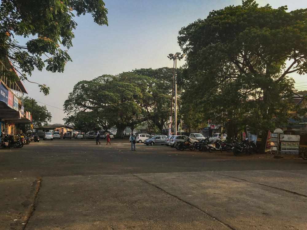 An open paved square in Vypin, Kerala, with large spreading trees lining the far side, where cars and rows of motorbikes are parked. A few pedestrians cross the empty foreground lot, with low shops and signboards along the left edge under a hazy late-afternoon sky.