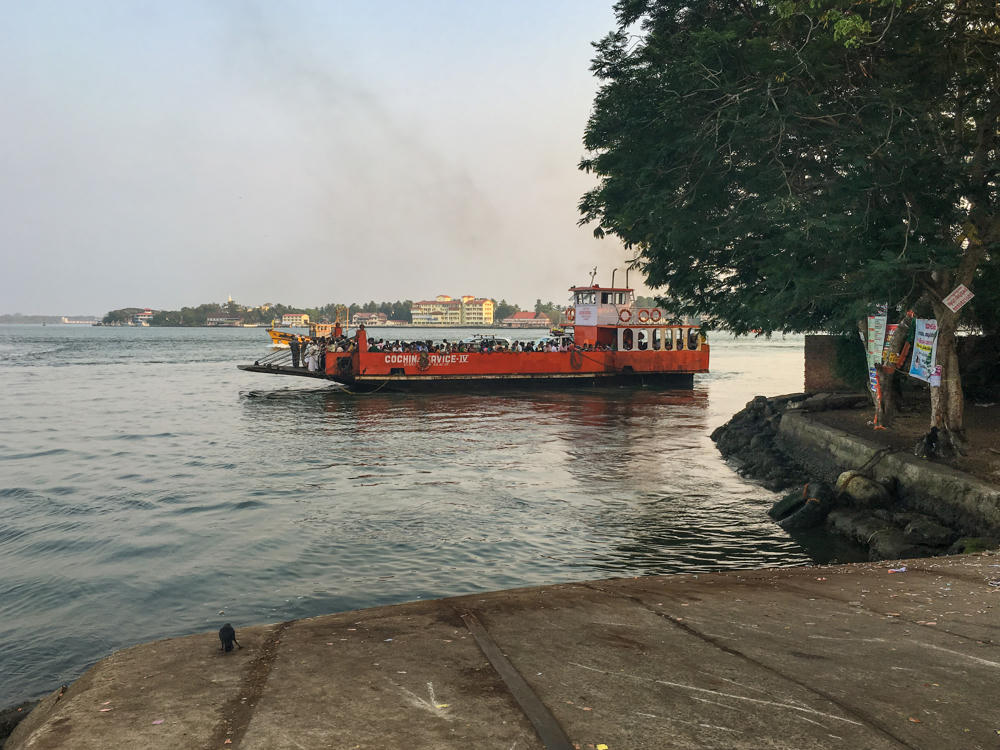 An orange ferry marked “Cochin Service IV” crowded with passengers crosses calm harbor water near Vypin, Kerala, with low buildings on the far shore under a hazy sky. In the foreground a concrete boat ramp leads to the water, and a large tree, posters, and a man in a white lungi stand along the embankment at right.