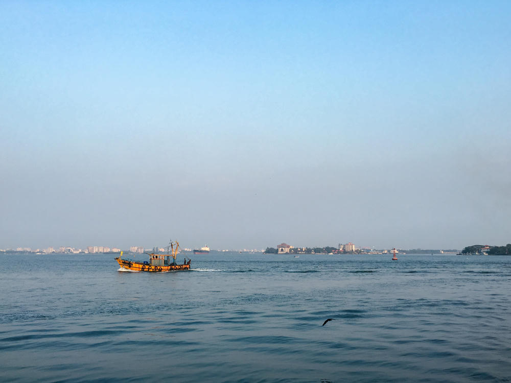 A yellow fishing boat moves across a wide expanse of calm blue-grey water under a hazy pale blue sky, with a distant low skyline of buildings on the horizon. A bird flies low over the water in the foreground, and a red navigation buoy floats to the right.