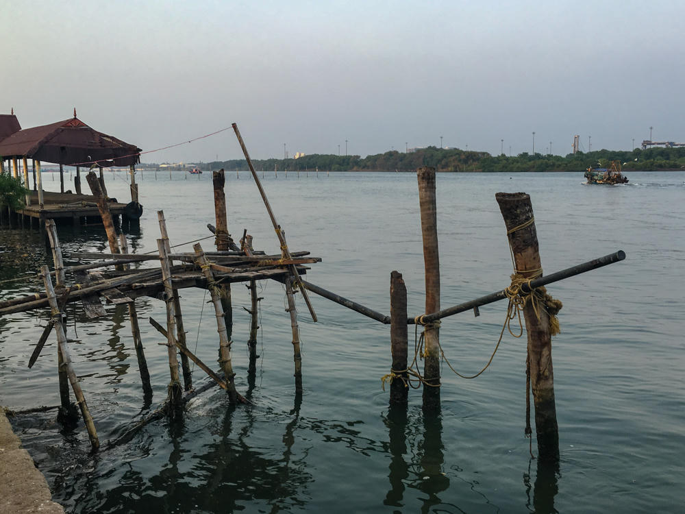 A weathered wooden and bamboo structure of poles tied together with rope stands in calm backwaters, with a tiled-roof pavilion on a jetty to the left and a low green shoreline with a fishing boat in the distance under a hazy pale sky at dusk.