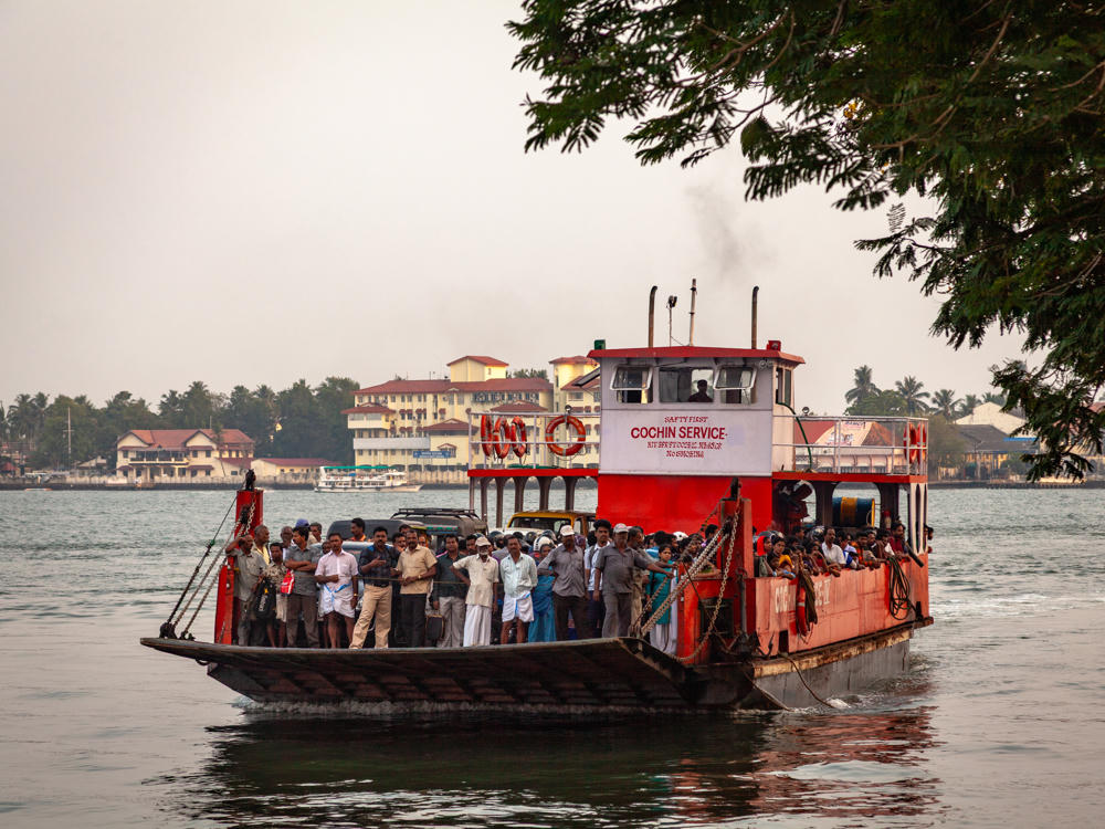 A crowded orange ferry marked “Cochin Service” carries dozens of passengers and several vehicles across calm water under a hazy grey sky, with a low town skyline of buildings and palm trees on the far shore and a tree branch overhanging from the upper right.