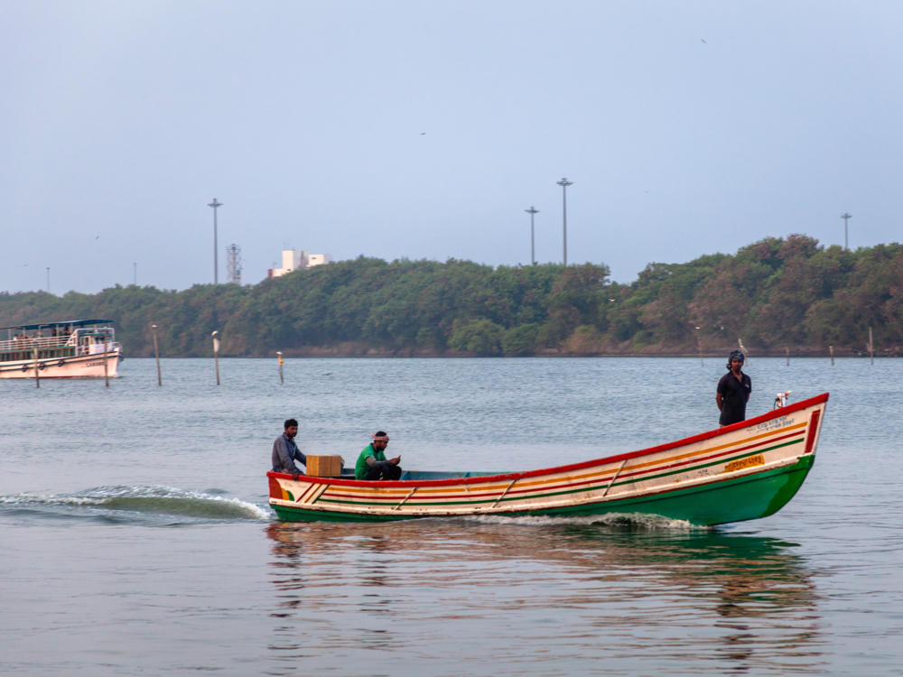 Three men ride in a small open boat painted green with red, yellow, and white stripes along its hull, motoring across calm water at Fort Vypin in Kerala, India. One man stands at the bow while two others sit near a cardboard box; a larger white passenger ferry and a tree-lined shore with tall light poles are visible in the hazy background.