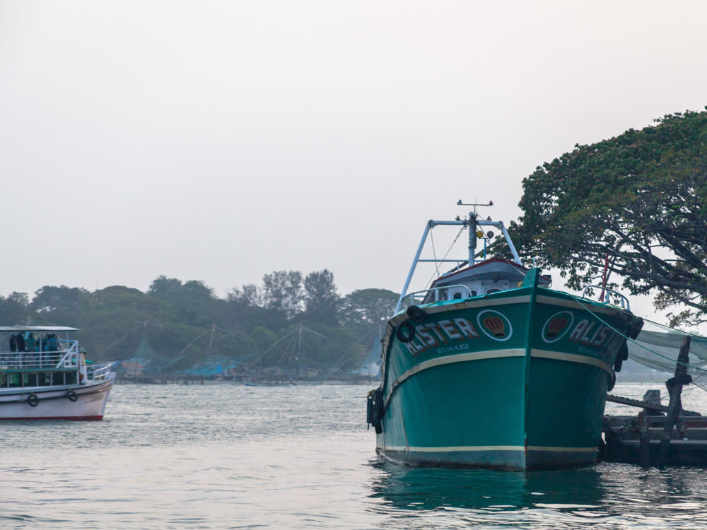 A green wooden fishing boat named “Alister” moored near the shore in calm water at dusk, with a large tree overhanging it on the right and Chinese fishing nets and a white passenger ferry visible in the hazy background.