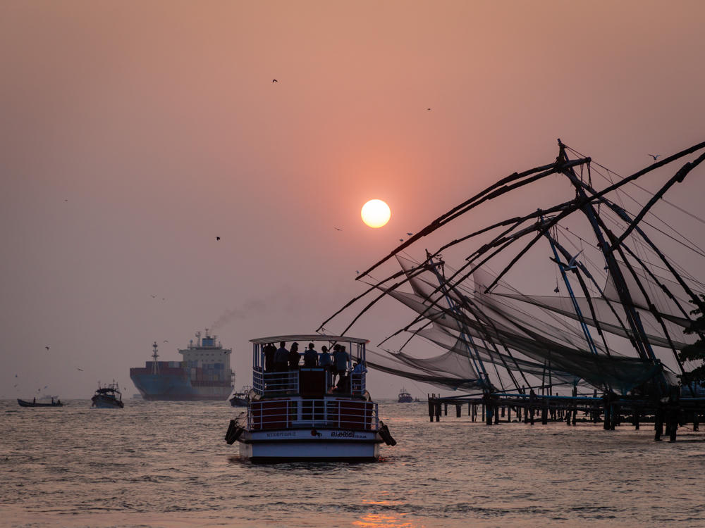 A passenger ferry crowded with people glides across hazy water at sunset, with the sun a soft pale disc low in the orange sky. On the right, traditional cantilevered Chinese fishing nets are silhouetted along the shore, while a large container ship and smaller fishing boats sit on the horizon.
