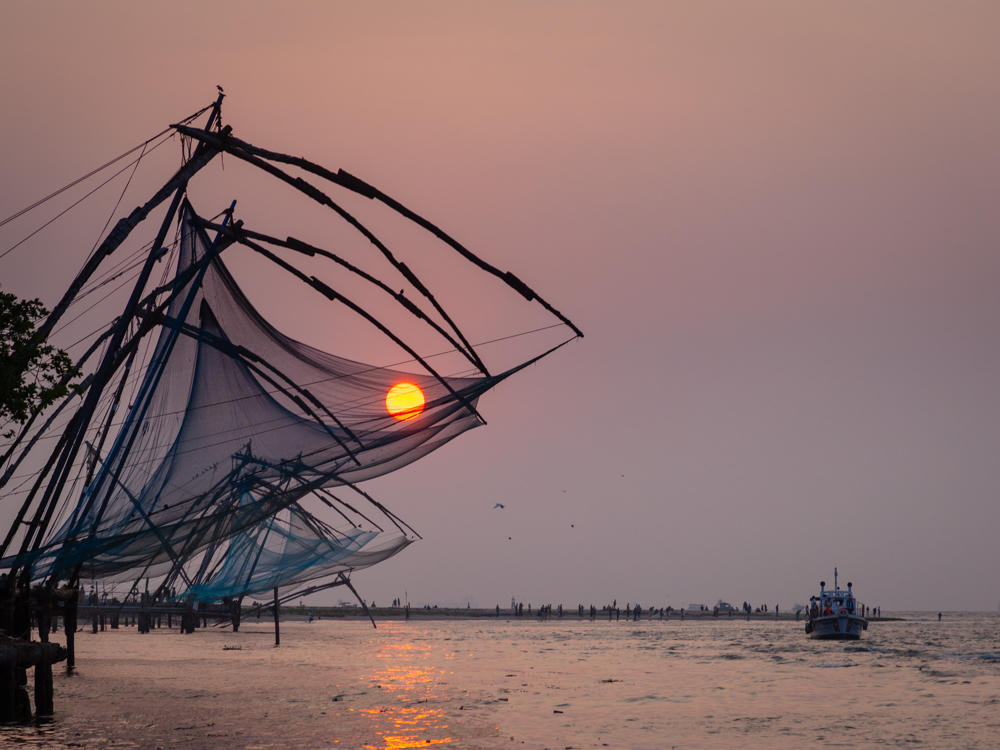 Cantilevered Chinese fishing nets silhouetted against a hazy pink sky at sunset, with the orange sun setting behind them over the water at Fort Kochi. Small boats and distant figures dot the calm sea on the right, and the sun’s reflection shimmers across the water.
