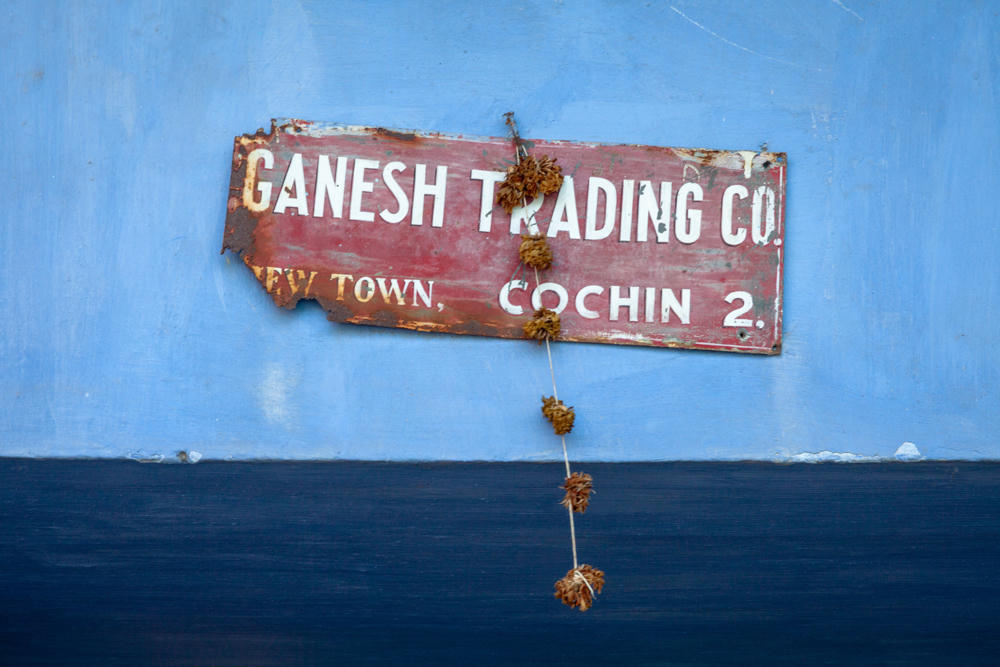 A weathered, rusty red metal sign reading “Ganesh Trading Co., New Town, Cochin 2.” in white lettering, mounted on a wall painted light blue above and dark navy below, with a string of dried marigold flowers hanging down over its surface.