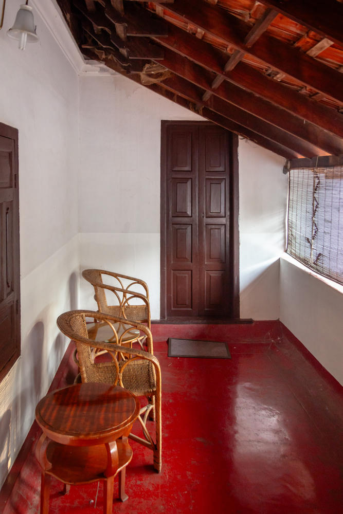 A narrow, traditional veranda with a polished red floor, whitewashed walls, and an exposed sloped wooden ceiling with rafters and clay tiles. Two woven wicker chairs and a small round wooden table sit along the left wall, with a tall dark paneled wooden door at the far end and a screened window letting in light on the right.
