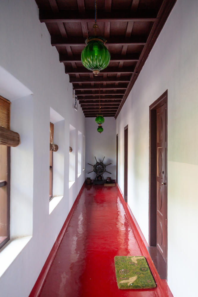 A long narrow corridor with a glossy deep-red floor, white plaster walls, and an exposed dark wooden beam ceiling, lit by green glass pendant lanterns hanging in a row. A wooden door stands on the right, bamboo blinds cover windows on the left, an antique ship’s wheel and pots sit at the far end, and a green floral mat lies on the floor in the foreground.