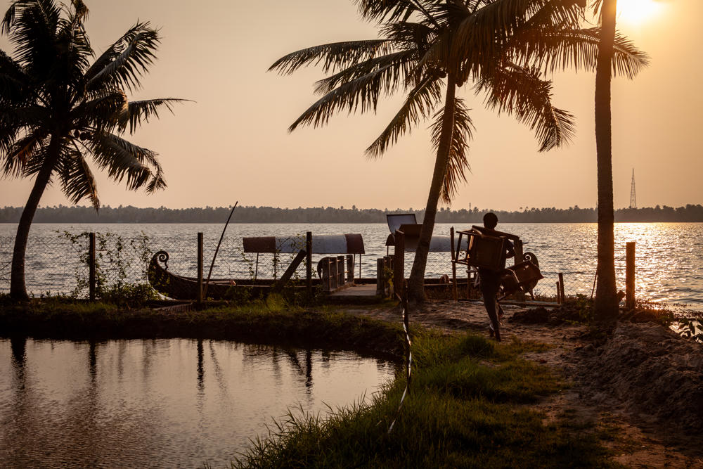A man carries stacked chairs along a sandy path between a backwater lagoon and a calm lake at golden hour, with tall coconut palms silhouetted against a bright hazy sky and a traditional carved wooden boat moored at a small jetty.