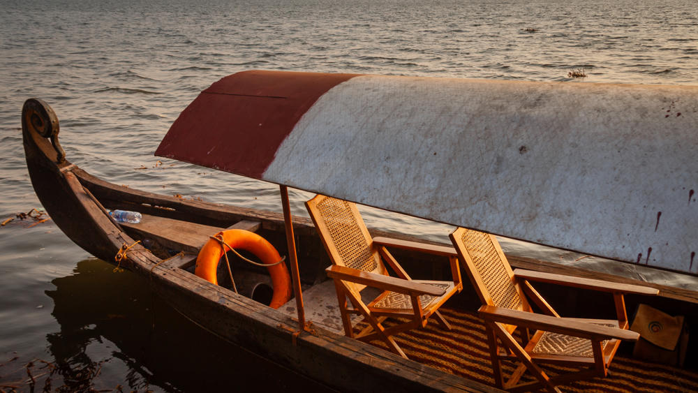 A traditional wooden canoe-style tour boat with a curved, decorated prow and an arched canopy floats on calm backwaters, fitted with two wooden folding chairs and an orange life ring, with a distant tree-lined shore on the horizon.