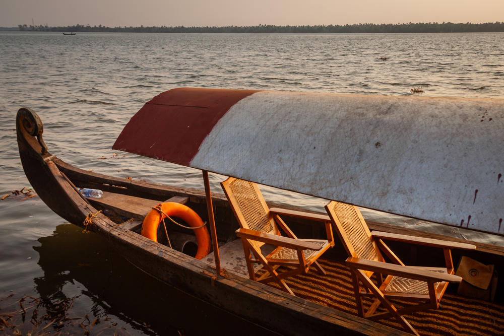 A traditional wooden canoe-style tour boat with a curved, decorated prow and an arched canopy floats on calm backwaters, fitted with two wooden folding chairs and an orange life ring, with a distant tree-lined shore on the horizon.