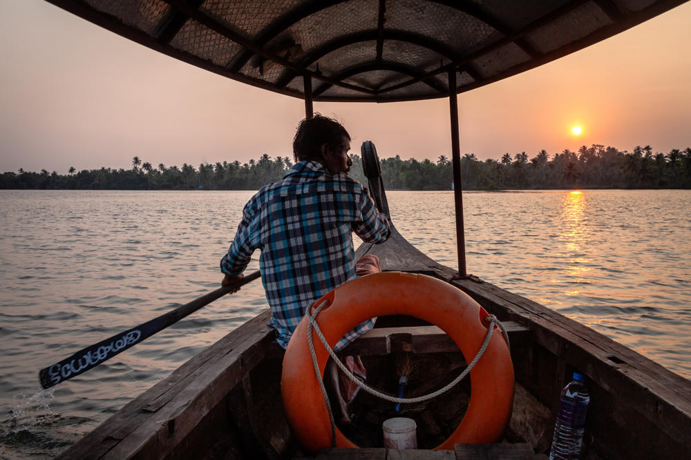 A man in a blue plaid shirt rows a wooden canopy boat with a hand-painted paddle across calm backwaters at sunset, viewed from inside the boat with an orange life ring and a water bottle in the foreground. Palm-fringed shoreline lines the horizon as the sun sinks low over the water.