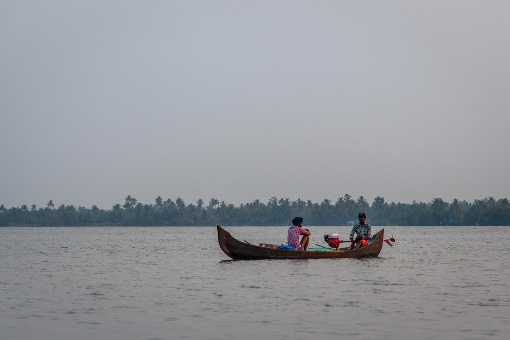 Two people sit in a small wooden canoe with a motor on calm, wide backwaters under a hazy pale sky, with a distant shoreline of palm trees on the horizon.